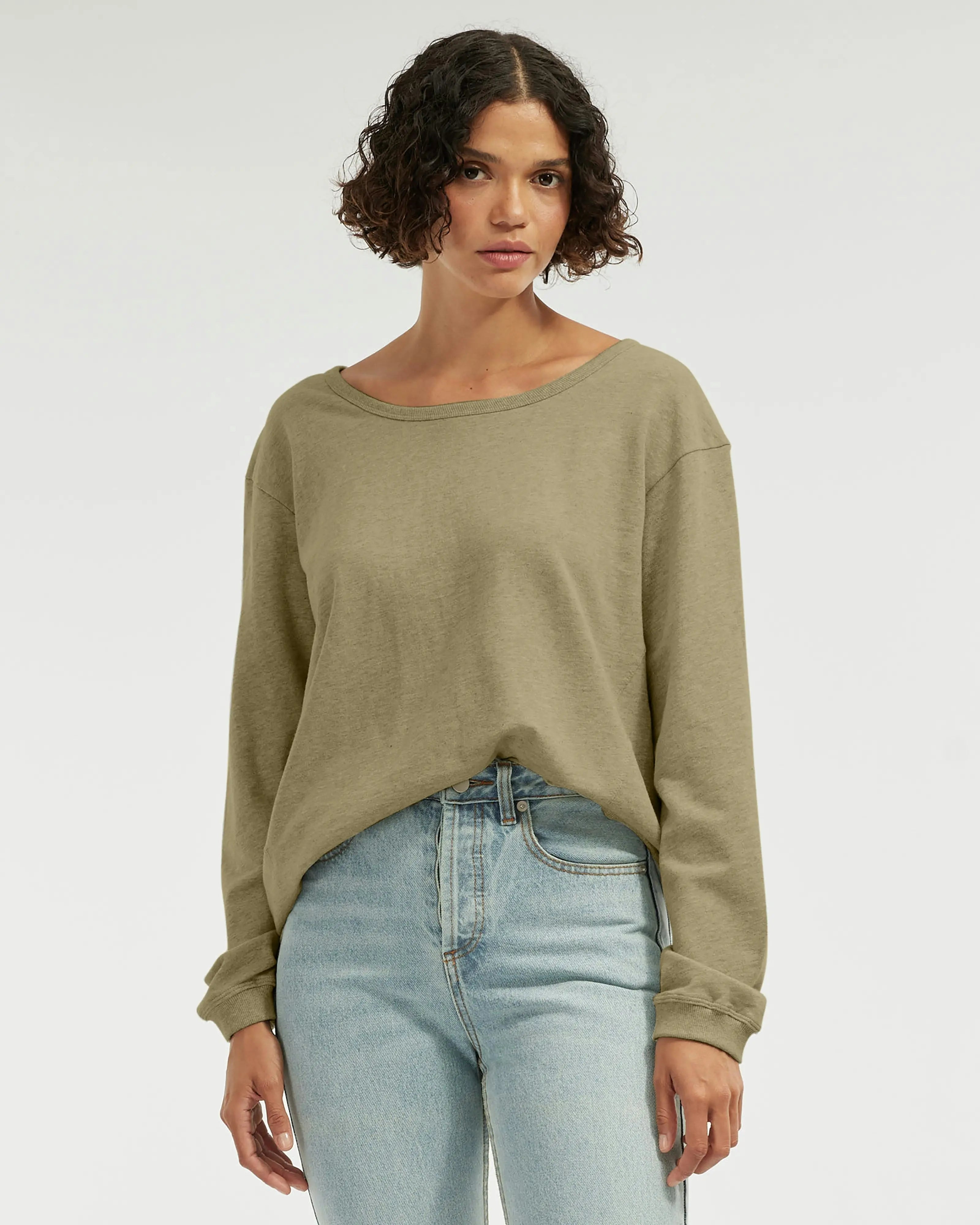 Model wearing a relaxed-fit long sleeve top in soft olive green, styled with light wash high-waisted jeans against a neutral background.
